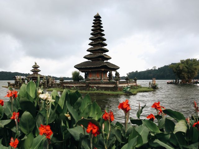 Ulun Danu Beratan multi-tier meru shrine on Lake Beratan under cloudy sky, red canna flowers in foreground