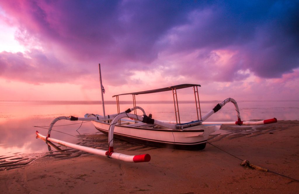 Traditional white outrigger jukung boat on a calm Bali beach under a colourful sunrise sky