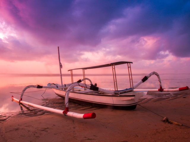 Traditional white outrigger jukung boat on a calm Bali beach under a colourful sunrise sky